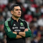 Entrenador de la Selección Mexicana con los brazos cruzados, ubicado del lado izquierdo de la imagen, durante un partido en un estadio con la afición desenfocada al fondo.