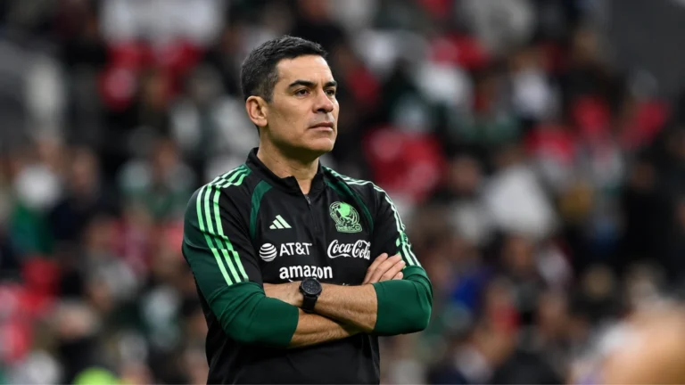 Entrenador de la Selección Mexicana con los brazos cruzados, ubicado del lado izquierdo de la imagen, durante un partido en un estadio con la afición desenfocada al fondo.